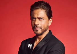 A well-dressed man in a black suit standing against a solid red background with a confident, serious expression.
