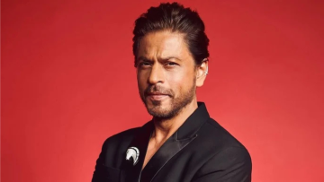 A well-dressed man in a black suit standing against a solid red background with a confident, serious expression.