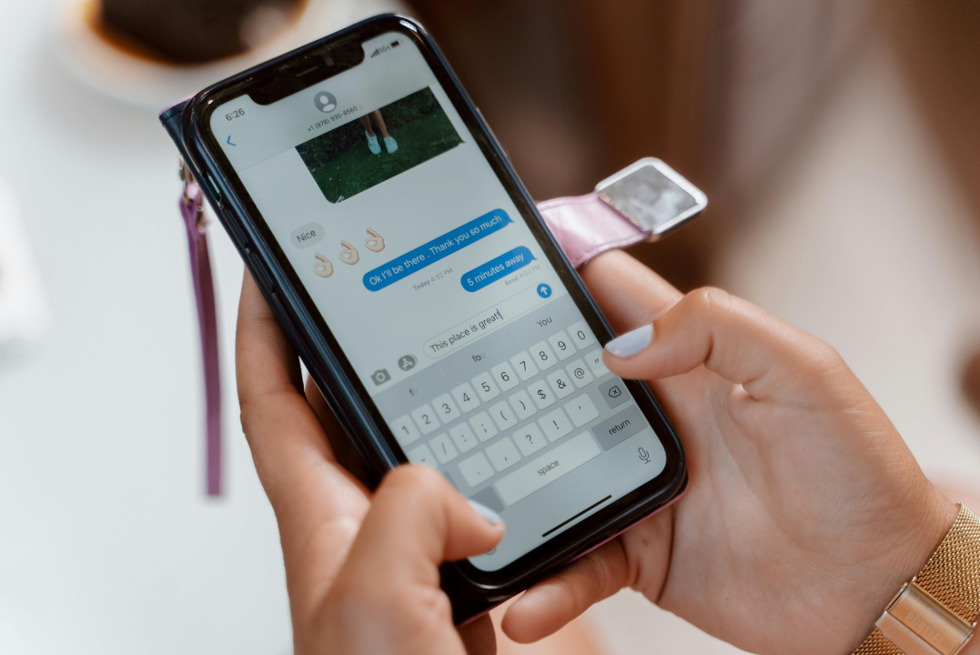 Close-up photo of a person holding a smartphone and typing a message on the screen, showing an active chat with text bubbles and emojis. The hands are visible with a gold wristwatch, and the phone is in a flip-style cover.