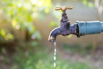 A close-up of an old outdoor water tap with a brass handle, dripping water against a blurred green background.