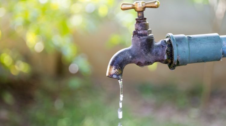 A close-up of an old outdoor water tap with a brass handle, dripping water against a blurred green background.