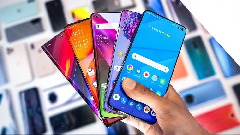 A hand holding four modern smartphones with colorful displays, shown against a blurred background of multiple mobile phones placed on a table.