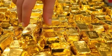 A close-up view of numerous shiny gold bars stacked together, with a hand reaching in to pick one up.