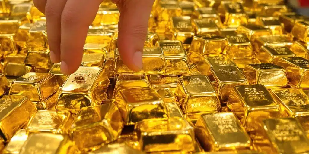 A close-up view of numerous shiny gold bars stacked together, with a hand reaching in to pick one up.
