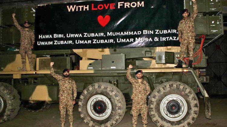 Pak Army ke chand faujis ek bade military vehicle par kharay hain. Vehicle ke upar ek bari black banner lagi hai jisper “With Love From” likha hua hai aur neeche kuch naam list kiye gaye hain. Faujis haath buland kar ke pose de rahe hain aur background aik andhera warehouse ya shed lag raha ha