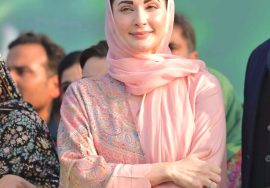 A female Pakistani political leader wearing a pink traditional dress and headscarf, standing confidently among people at a public gathering
