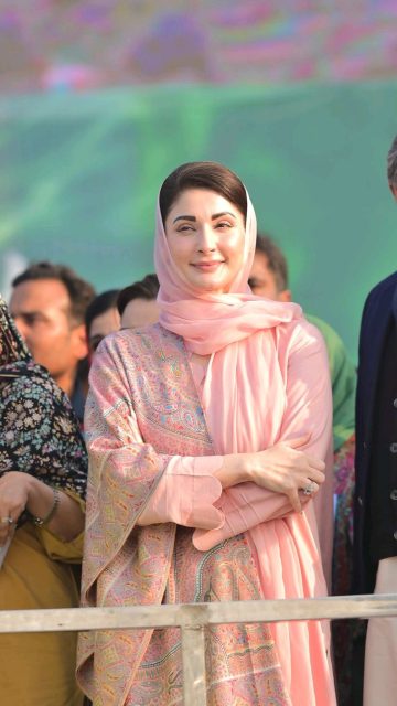 A female Pakistani political leader wearing a pink traditional dress and headscarf, standing confidently among people at a public gathering