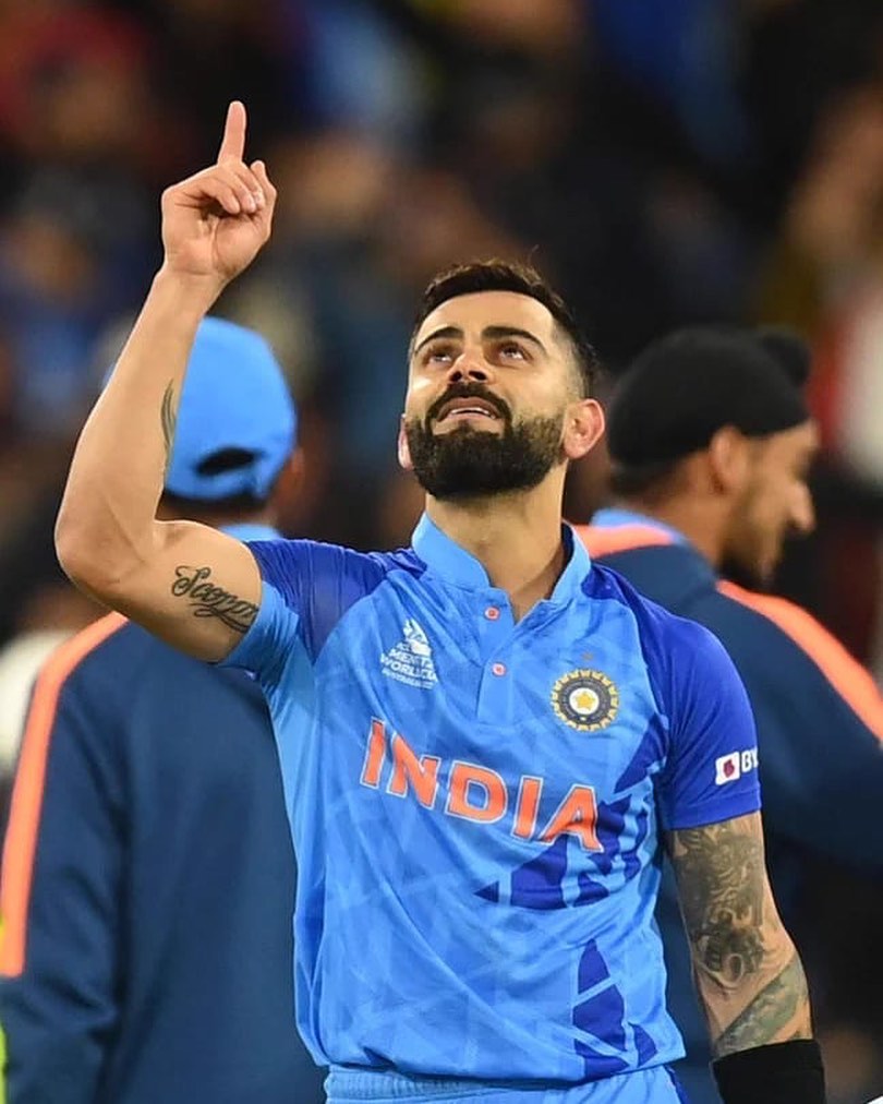 An Indian cricketer in a blue India jersey raises one finger toward the sky while looking upward, celebrating on the field.
