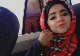 A young woman wearing a red and black checkered scarf takes a selfie inside a vehicle, gently holding her scarf near her chin. Soft light comes through the window behind her.