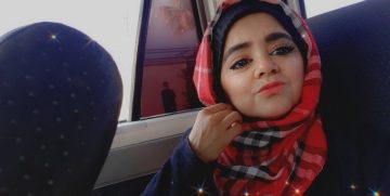 A young woman wearing a red and black checkered scarf takes a selfie inside a vehicle, gently holding her scarf near her chin. Soft light comes through the window behind her.