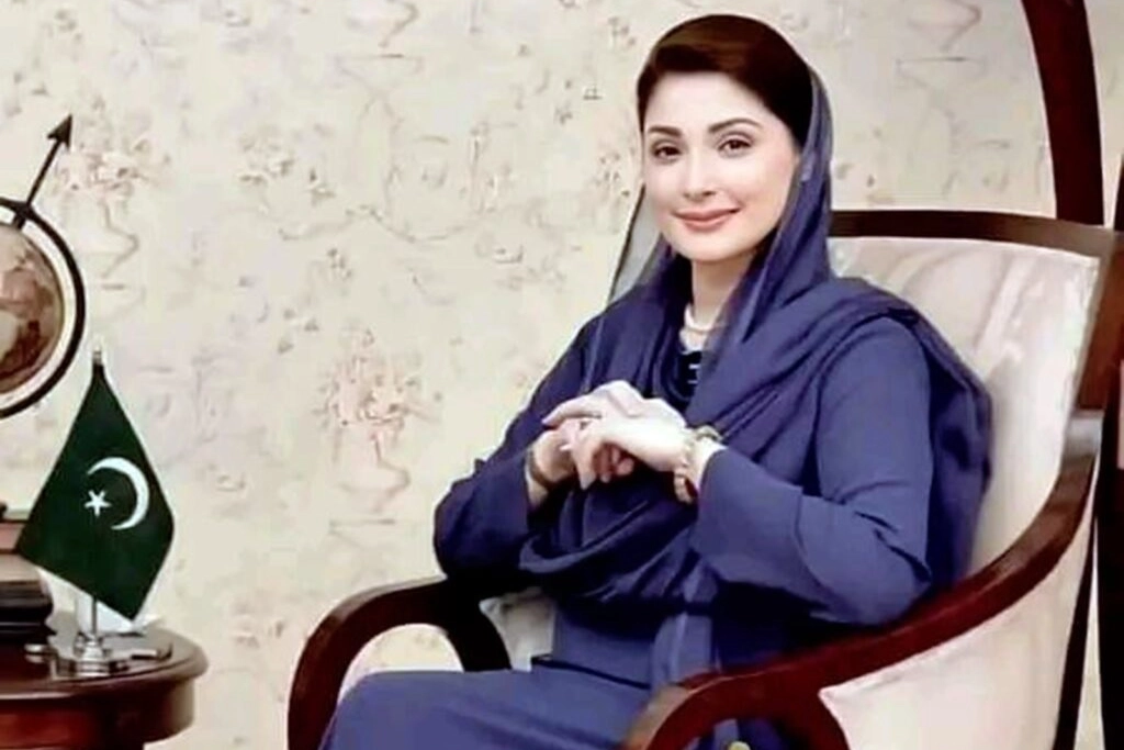 A woman wearing a blue traditional outfit and headscarf sits on a wooden chair, smiling softly. A small table with a globe and a Pakistani flag is placed beside her.