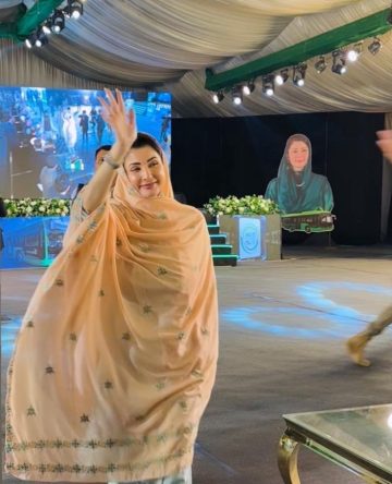 A woman in a peach embroidered dupatta and light blue outfit is smiling and waving while walking inside a decorated event hall with bright stage lights, floral arrangements, and a large portrait of herself displayed in the background.