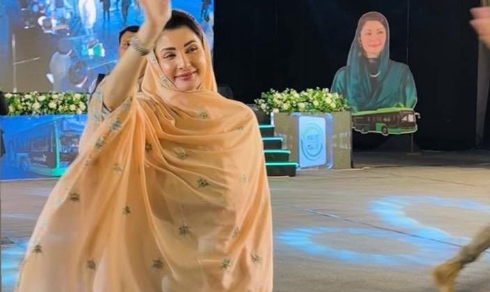 A woman in a peach embroidered dupatta and light blue outfit is smiling and waving while walking inside a decorated event hall with bright stage lights, floral arrangements, and a large portrait of herself displayed in the background.