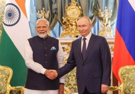 Two formally dressed men stand in an ornate room and shake hands in front of large national flags. The setting includes gold-decorated furniture, chandeliers, and an official backdrop.