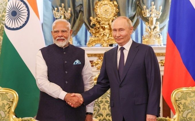 Two formally dressed men stand in an ornate room and shake hands in front of large national flags. The setting includes gold-decorated furniture, chandeliers, and an official backdrop.