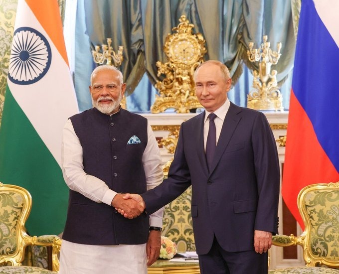 Two formally dressed men stand in an ornate room and shake hands in front of large national flags. The setting includes gold-decorated furniture, chandeliers, and an official backdrop.