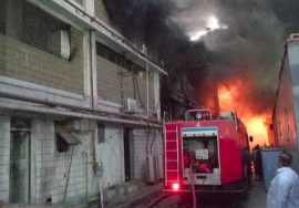 A large industrial building is engulfed in heavy smoke and flames while a red fire truck and firefighters work to control the blaze. Thick black smoke rises into the sky as bright orange fire burns inside the structure.