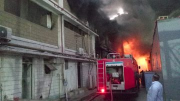 A large industrial building is engulfed in heavy smoke and flames while a red fire truck and firefighters work to control the blaze. Thick black smoke rises into the sky as bright orange fire burns inside the structure.