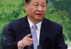 "A man in a formal suit speaking during a meeting, seated at a table with microphones in front of him."