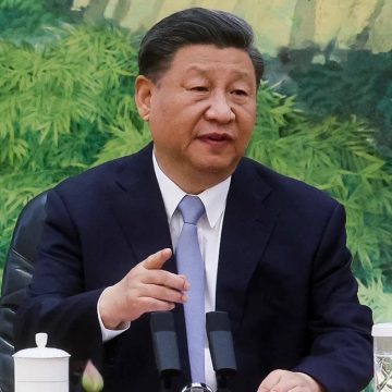 "A man in a formal suit speaking during a meeting, seated at a table with microphones in front of him."