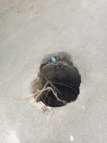 An open and broken manhole in the middle of a road, posing a serious danger to pedestrians, children, and vehicles.