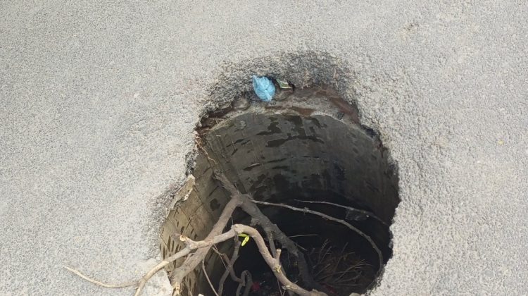 An open and broken manhole in the middle of a road, posing a serious danger to pedestrians, children, and vehicles.