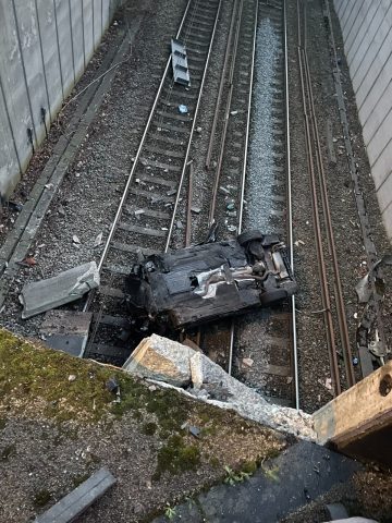 An overturned and wrecked car lying on railway tracks after crashing from a height, with debris scattered across the train line.