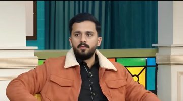 "A man sitting indoors wearing a brown jacket with a shearling collar during a studio interview setting."