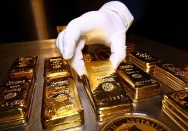 A gloved hand carefully lifting a gold bar from a table filled with multiple stacked gold bullion bars.