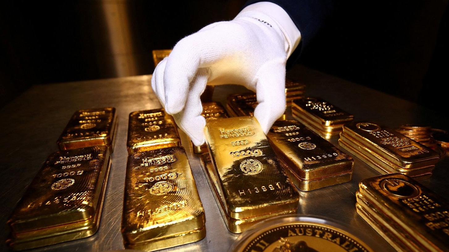 A gloved hand carefully lifting a gold bar from a table filled with multiple stacked gold bullion bars.