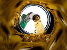 A close-up view of golden fuel being poured from a green fuel nozzle into a container, captured from inside the container with liquid swirling around the edges.