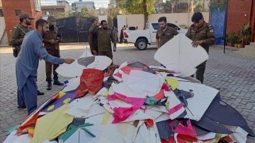 A group of Pakistani police officers stand around a large pile of confiscated kites and string on a paved courtyard, inspecting them as part of a crackdown on kite flying.