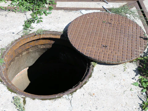Open manhole with a partially lifted metal cover on a concrete surface, showing a dark underground opening.